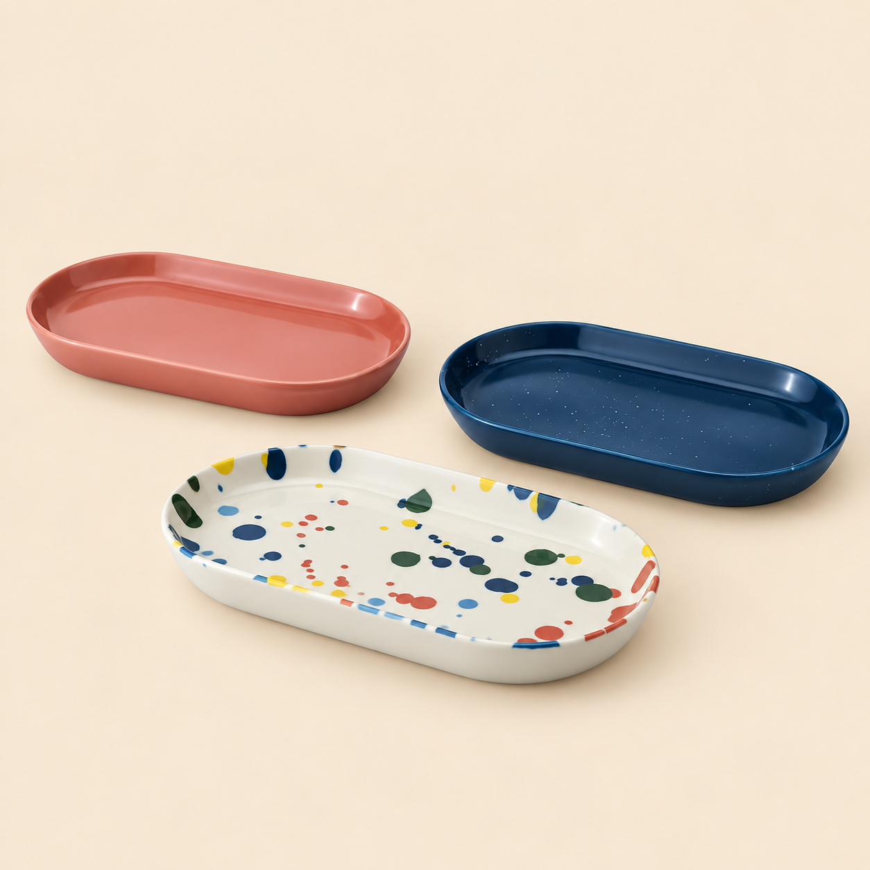 Three LIVA oval stoneware serving plates shown together, including solid pink, solid deep blue, and white with multi-colored paint splatter designs against a neutral background.